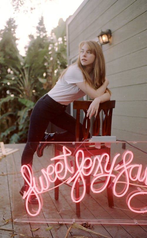 Confident young blonde in jeans and white t shirt leaning on chair and looking away against pink neon sign