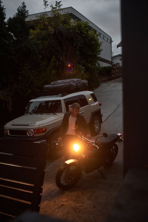 From above of anonymous young male biker in leather jacket parking motorcycle near SUV car on empty road