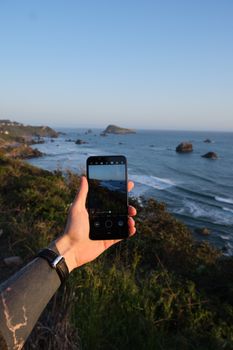 Crop faceless young tattooed male traveler taking photo of sunset over sea on smartphone during summer holidays
