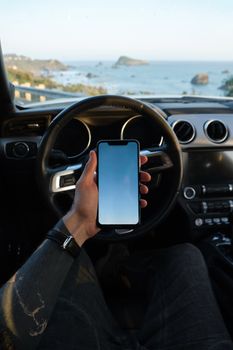 From above of crop unrecognizable tattooed male traveler using app on smartphone while sitting on driver seat in modern car against blurred seascape