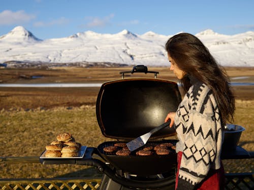 Free Side view of young female in warm sweater holding spatula and grilling meat while preparing food during picnic in countryside Stock Photo