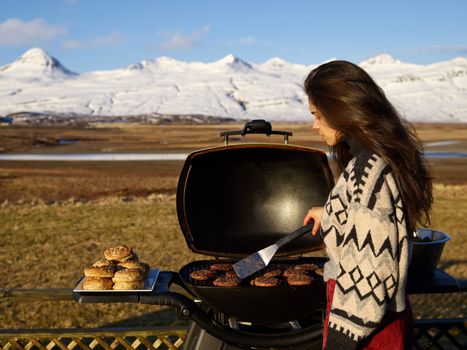 Side view of young female in warm sweater holding spatula and grilling meat while preparing food during picnic in countryside