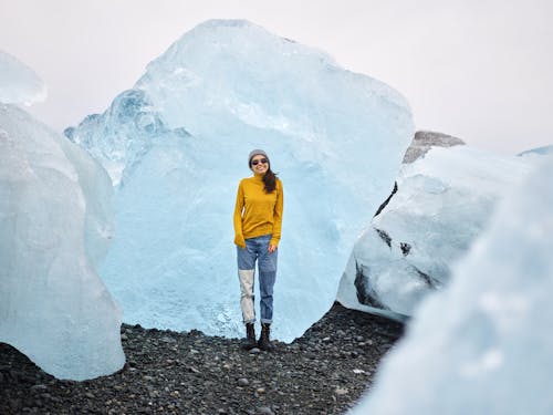 Cheerful young woman in sunglasses standing on rocky ground among multicolored huge ice blocks during trip to north