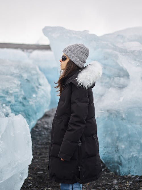 Back view of woman in warm outerwear and sunglasses standing among frozen blocks and enjoying ice landscape during vacation in frozen country