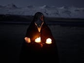 一个戴着口罩、身份不明的人，在夜晚的雪山前手持燃烧的蜡烛，凝视着镜头