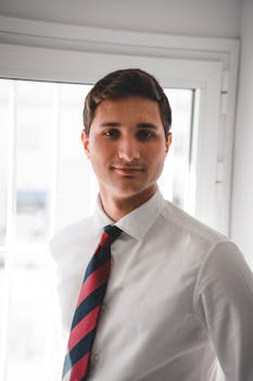 Portrait of a confident young man in a white shirt and striped tie, standing near a window.