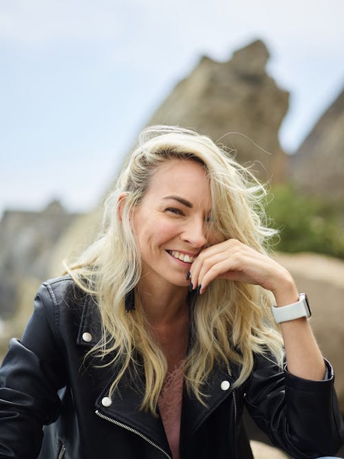 From below of young positive lady with blond hair in leather jacket smiling and touching face while looking at camera on sunny day