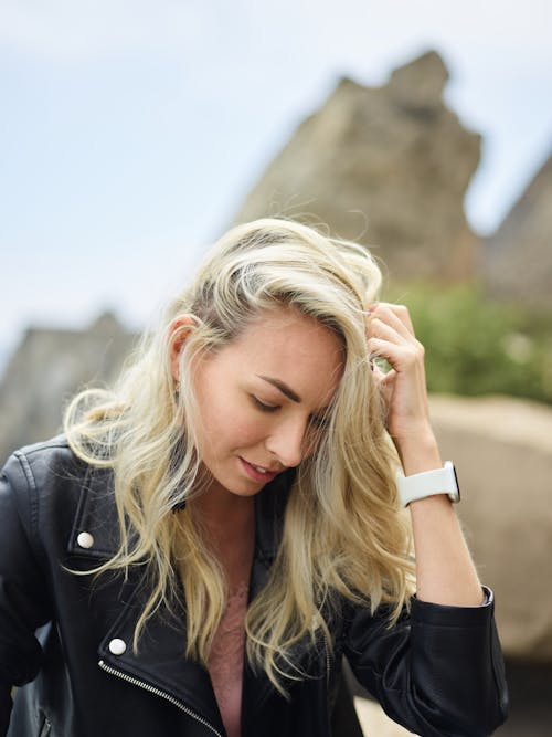 Peaceful young female in leather jacket and smartwatch looking down and touching hair against blurred cliff on sunny day