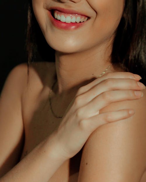 Crop young female with perfect skin covering breast and smiling brightly in soft light