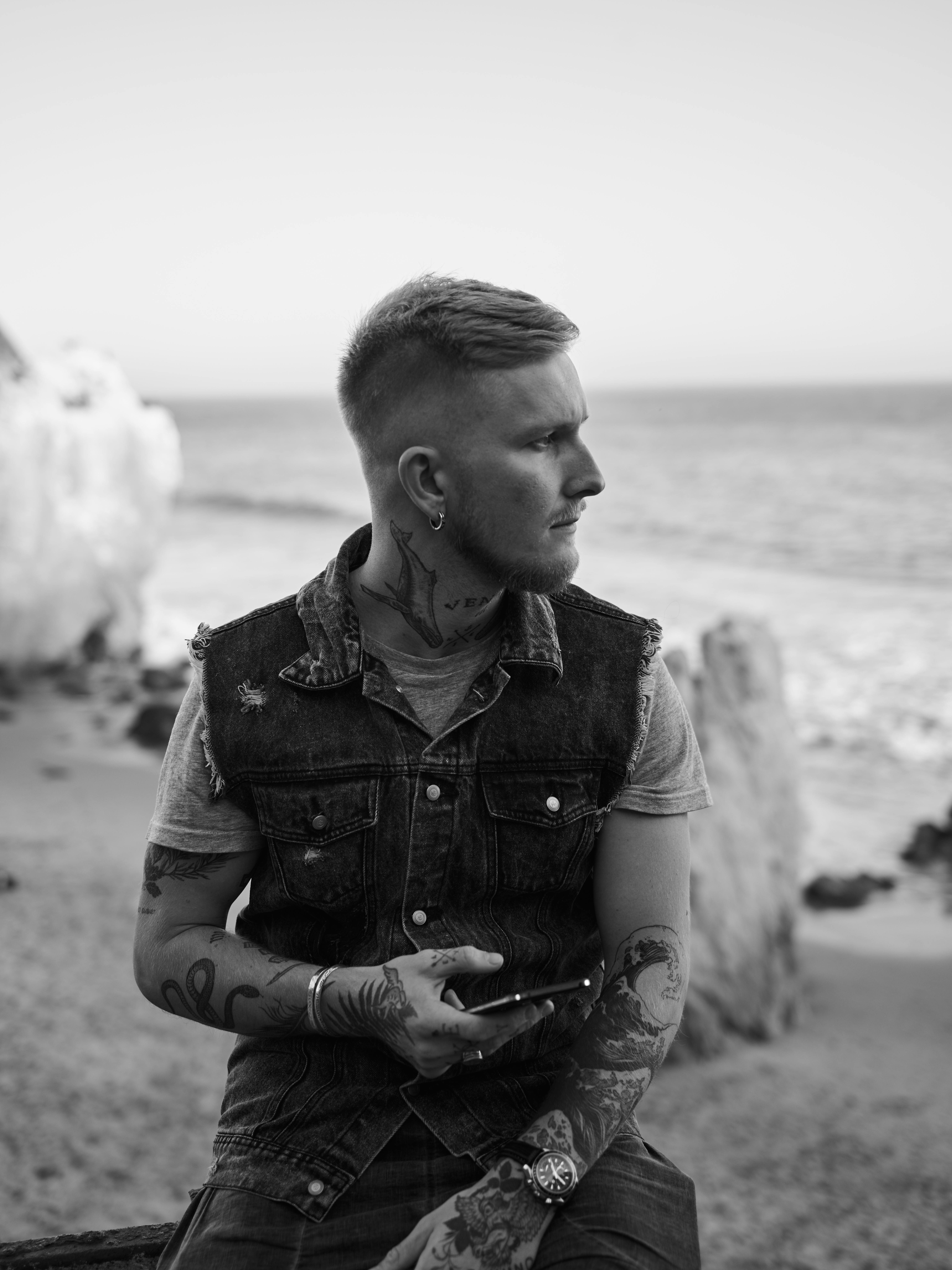 Serious young tattooed man with smartphone in stylish outfit relaxing on sandy beach of rocky ocean and looking away
