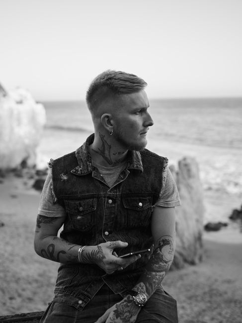 Serious young tattooed man with smartphone in stylish outfit relaxing on sandy beach of rocky ocean and looking away