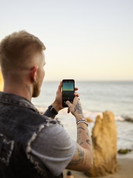 Back view of anonymous young tattooed male traveler taking photo of rocky seashore during summer holidays