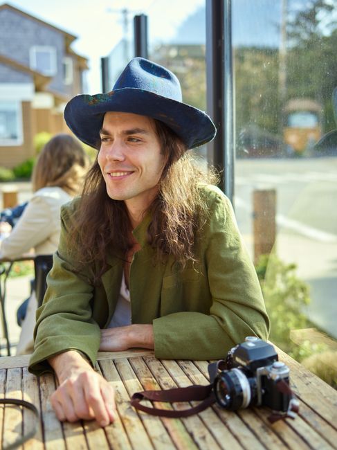 Positive young stylish man with long hair in trendy hat siting at wooden table with photo camera while resting in outdoor cafe