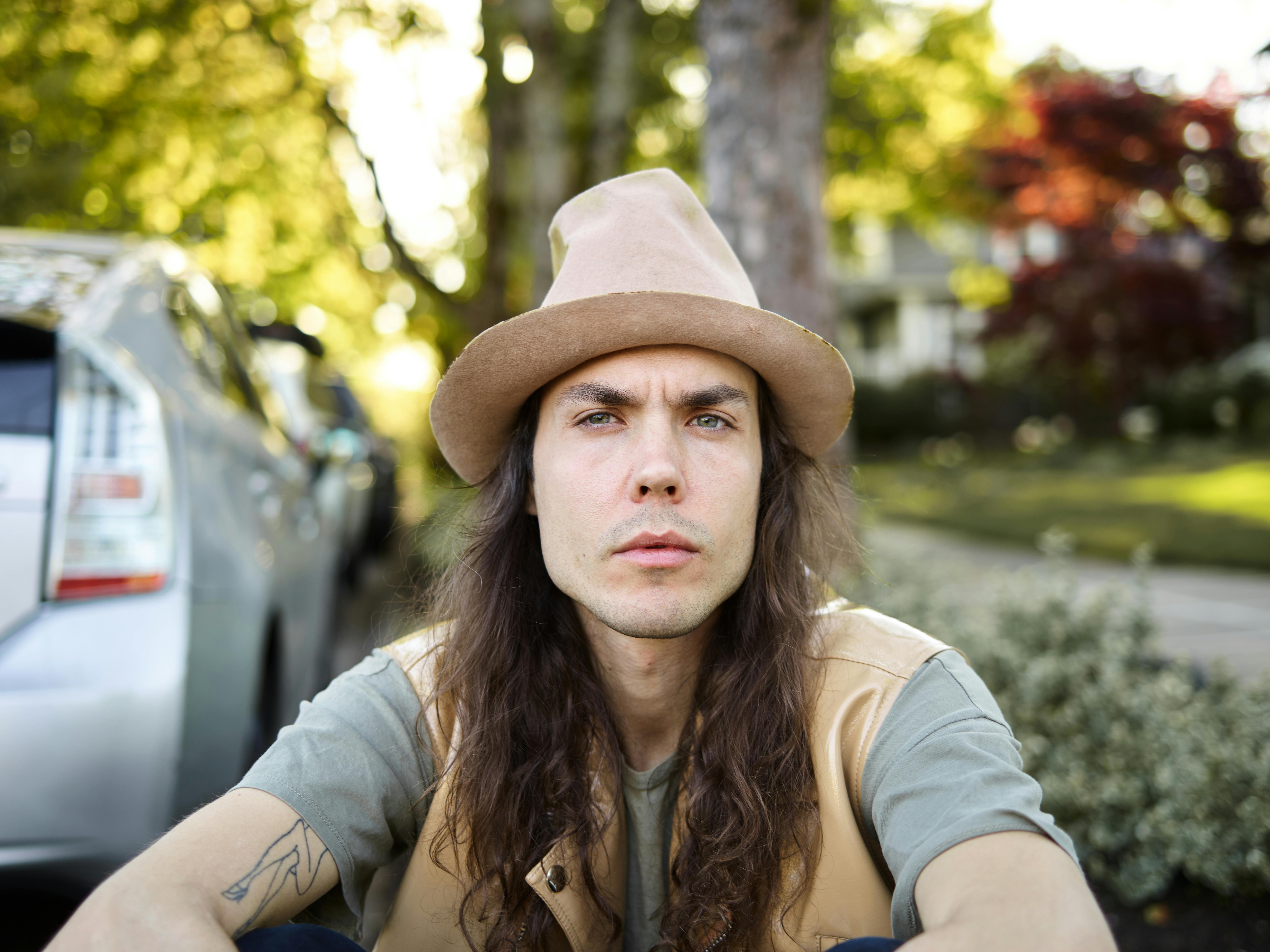 Slender serious long haired male in hat sitting under tree and frowning while looking at camera on street in sunny day