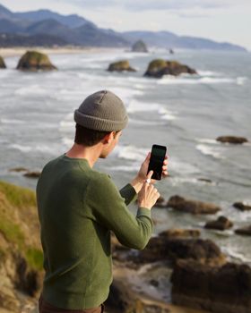 Back view of male tourist taking photo of sea and rocks on smartphone while exploring rocky coast