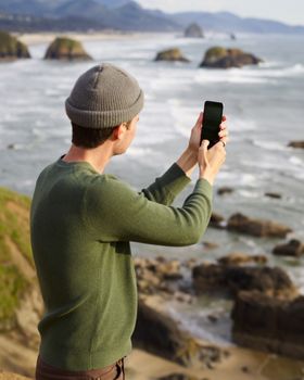 Side view of unrecognizable male traveler in hat and jumper looking at seascape and taking photo of vast sea surface on mobile phone