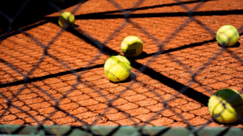 Close-up view of tennis balls scattered on a clay court, captured through a chain-link fence.