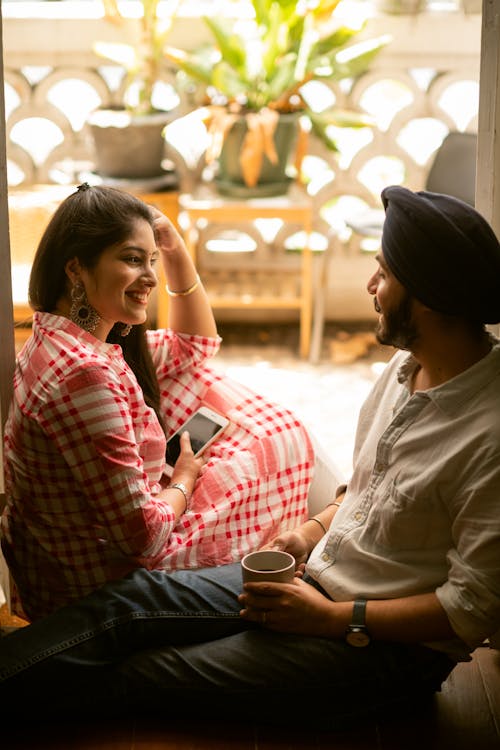 Side view of young Indian man with cup of coffee and woman with smartphone in hand smiling and talking happily while sitting on threshold of balcony