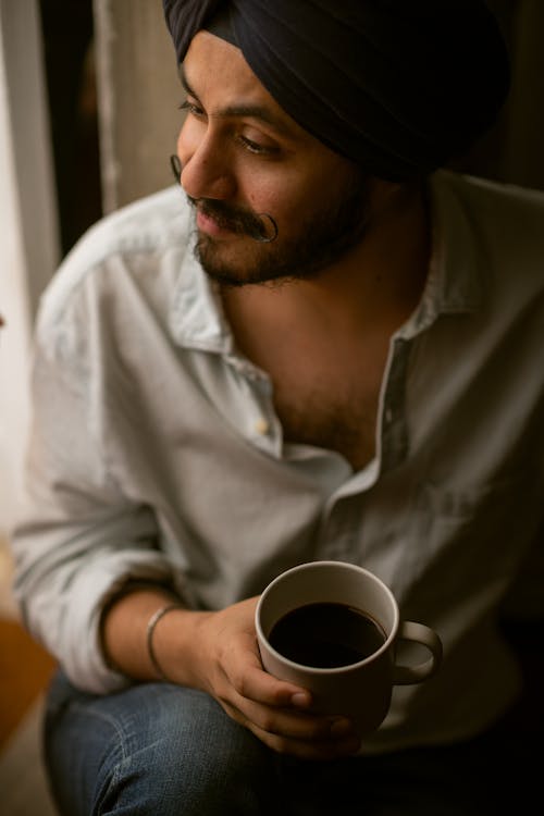 From above of positive young Indian male with handlebars in turban and casual shirt undone at collar sitting on floor with cup of black coffee in hand and looking away