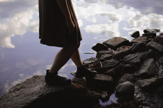 Side view of crop anonymous female in dress and boots stepping on stony coast near tranquil lake water with reflection of cloudy sky