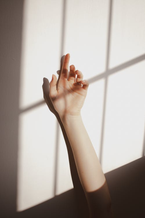 Crop anonymous female keeping raised hand against white wall with sunlight and shadow of window grate