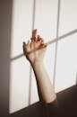 Crop anonymous female keeping raised hand against white wall with sunlight and shadow of window grate