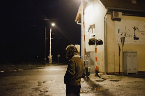 Side view of lonely unrecognizable male in warm jacket standing near illuminated railway crossing with shabby building and barriers at night time