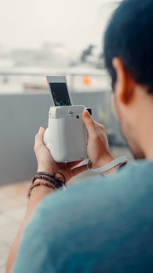 Back view of crop unrecognizable male traveler in casual clothes taking picture with instant photo camera while standing against blurred background