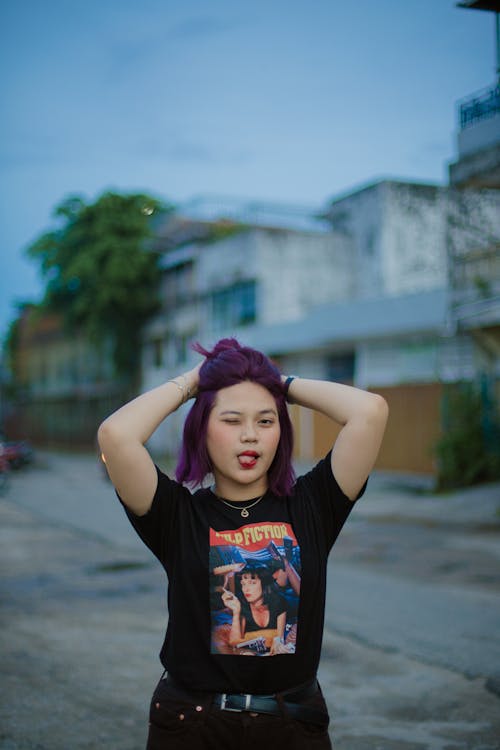 Free Asian teen student with purple dyed hair wearing trendy t shirt with modern print showing tongue and looking at camera while standing against blurred urban street Stock Photo