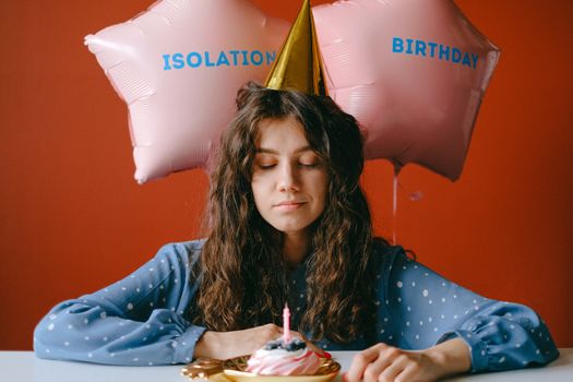 A woman in a party hat celebrates her birthday in isolation with balloons and a cupcake.