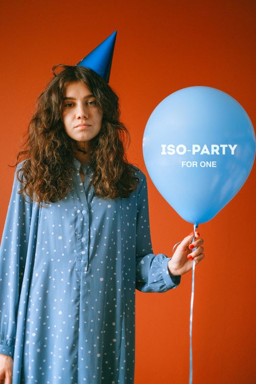 Free Young woman in a blue dress holds a balloon with 'ISO-PARTY FOR ONE' text against an orange background. Stock Photo
