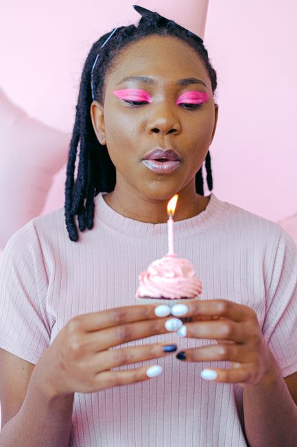 African American woman making a birthday wish, blowing candle on cupcake with a pink background.