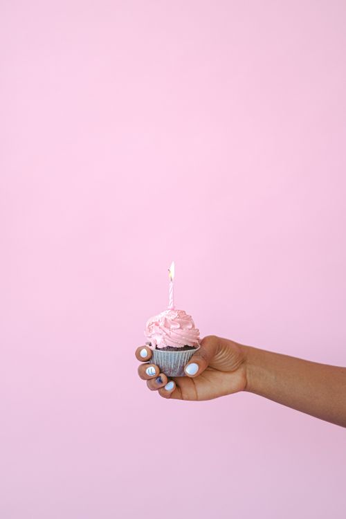 Free A cupcake with a lit candle held in a hand against a pastel pink background. Stock Photo