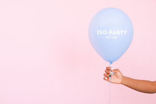Free A hand holding a blue balloon with 'ISO-PARTY FOR ONE' text on a light pink background. Stock Photo
