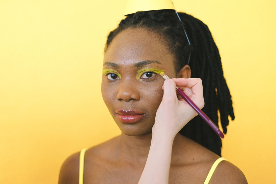 Close-up of an African American woman with dreadlocks receiving a makeup touch-up against a yellow background.