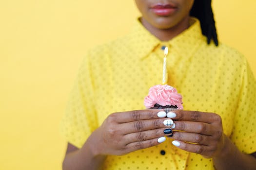 Close-up of a woman holding a cupcake with a lit candle against a bright yellow background.