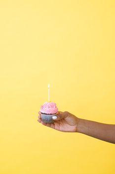 Colorful cupcake with lit candle held against a vibrant yellow backdrop, ideal for celebrations.