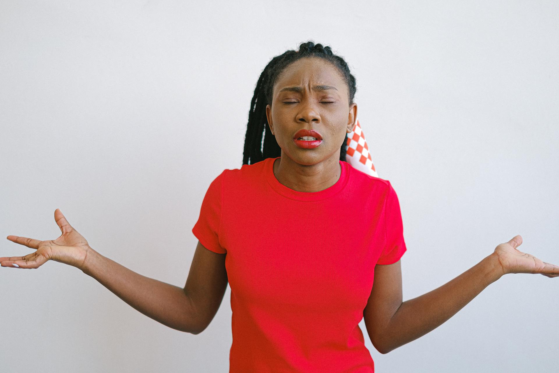 Portrait of a woman in a red shirt expressing confusion with her eyes closed and arms out. Ideal for emotion concepts.