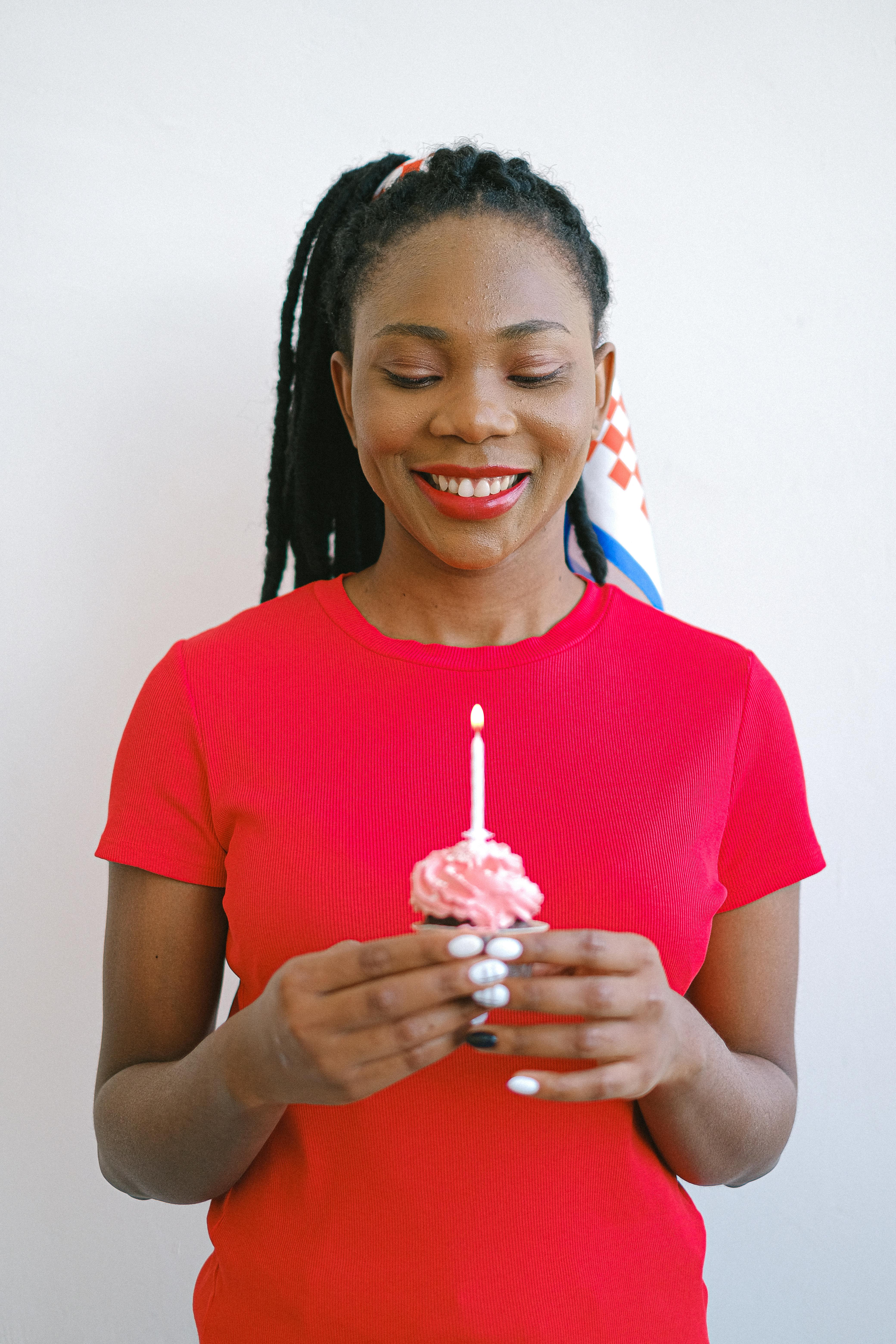 A joyful woman in a red shirt holding a candlelit cupcake, symbolizing celebration.