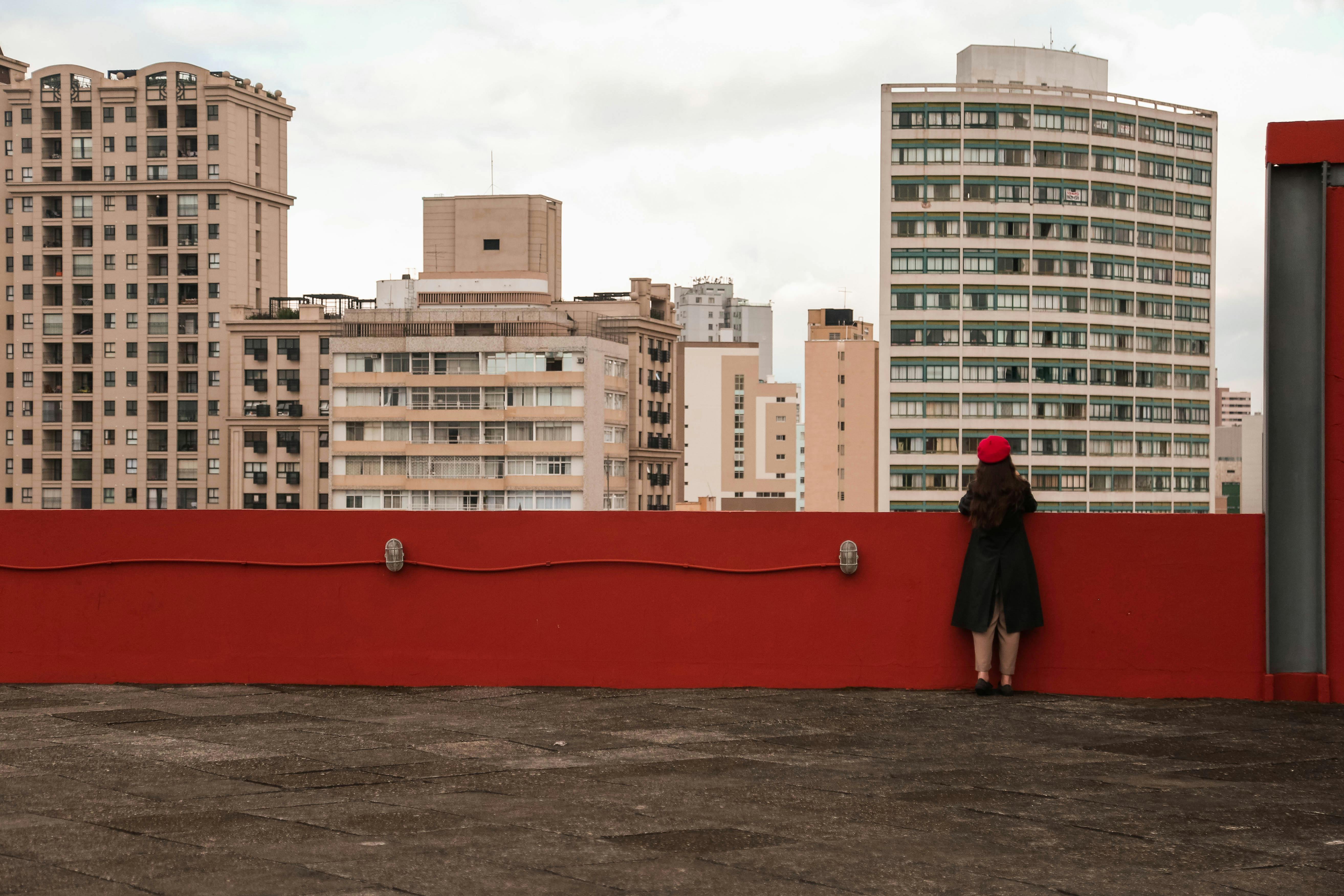 Back view of anonymous female in outerwear contemplating contemporary multistory residential buildings while standing on shabby fenced roof in city