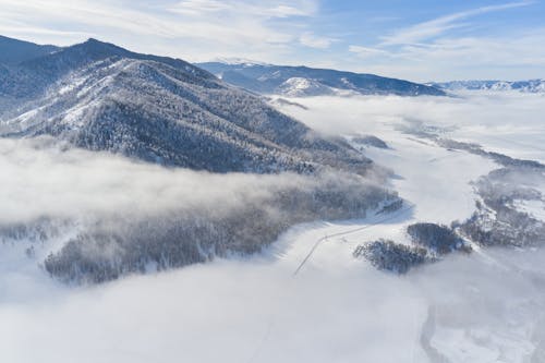 Drone view of snowy valley among mountains covered coniferous forest and road going between mountain ridge through clouds in sunny winter day