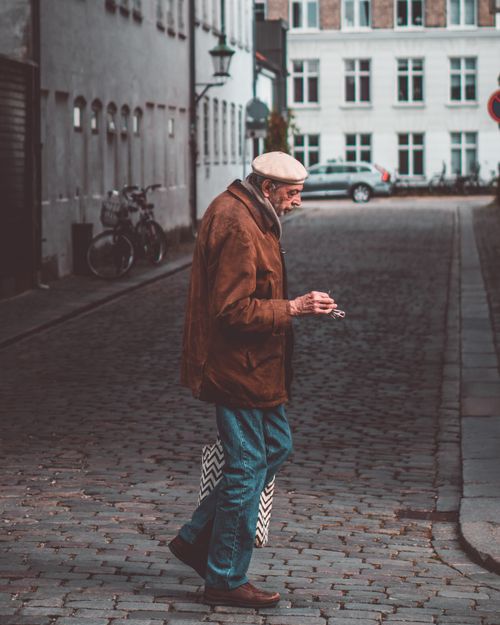 Free From above side view of senior male in outerwear with bag strolling on pavement between old houses in town Stock Photo