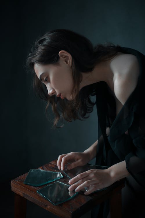 Free From above side view of young sad ethnic female leaning forward while touching broken mirror pieces in dark room Stock Photo