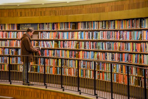 Side view of anonymous male standing near bookshelves with collection of colorful books in city library