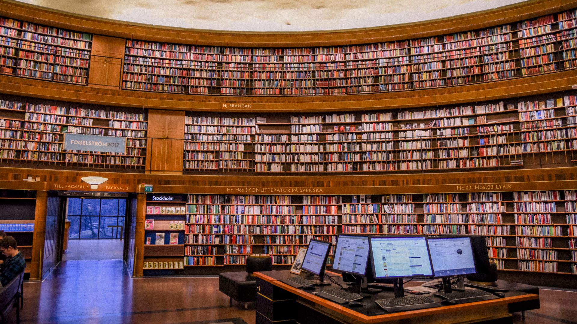 Public library with collection of colorful books on wooden bookshelves near table with computer monitors
