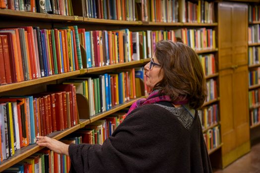 Side view of female looking for book while standing near bookshelves with multicolored covers of literature collection in city library