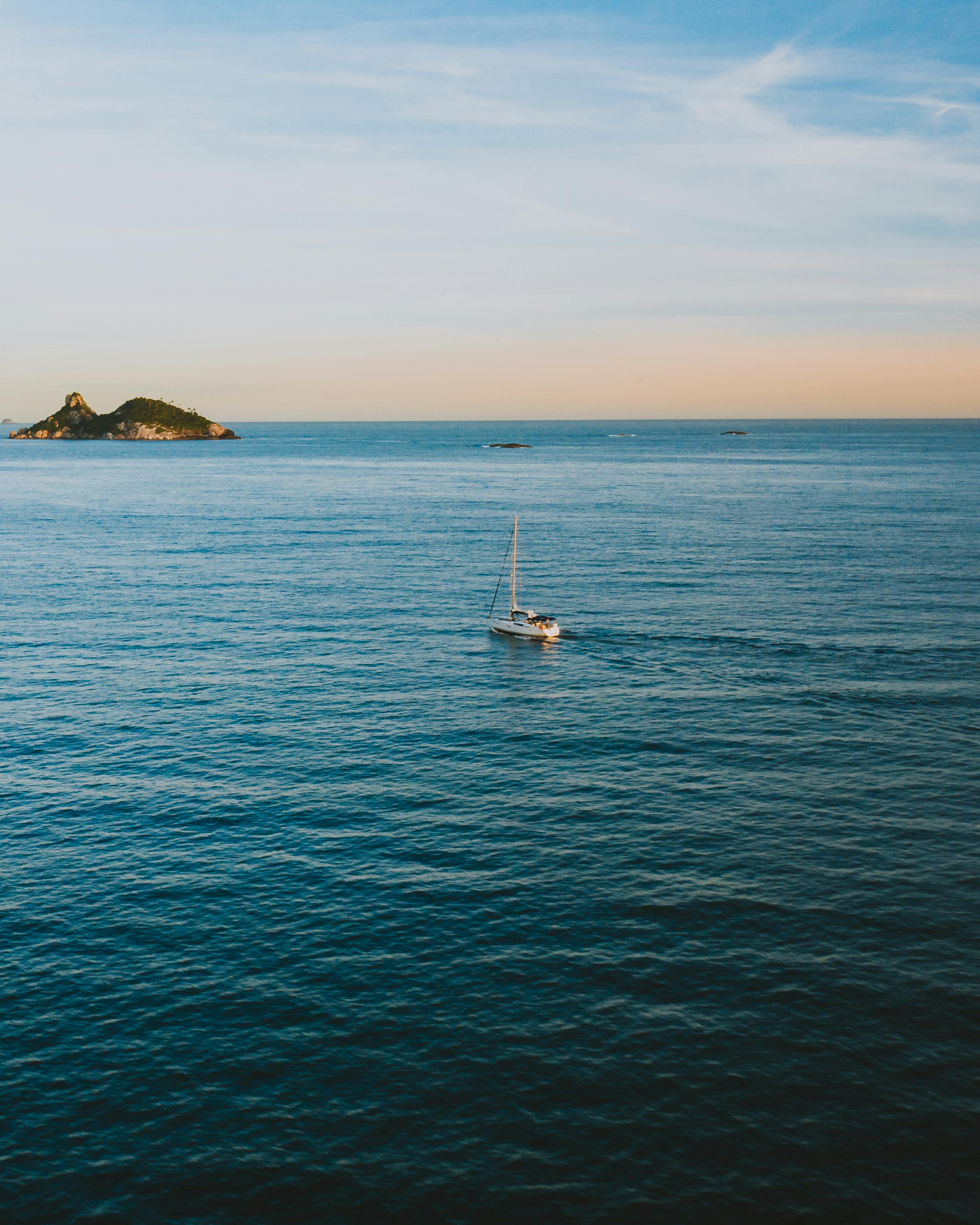 A serene sailboat drifts on the open ocean with a distant island backdrop, captured from above.