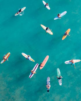 Photo by Jess Loiterton A vibrant aerial shot of surfers on colorful boards in turquoise waters, capturing Hawaiian surfing culture.