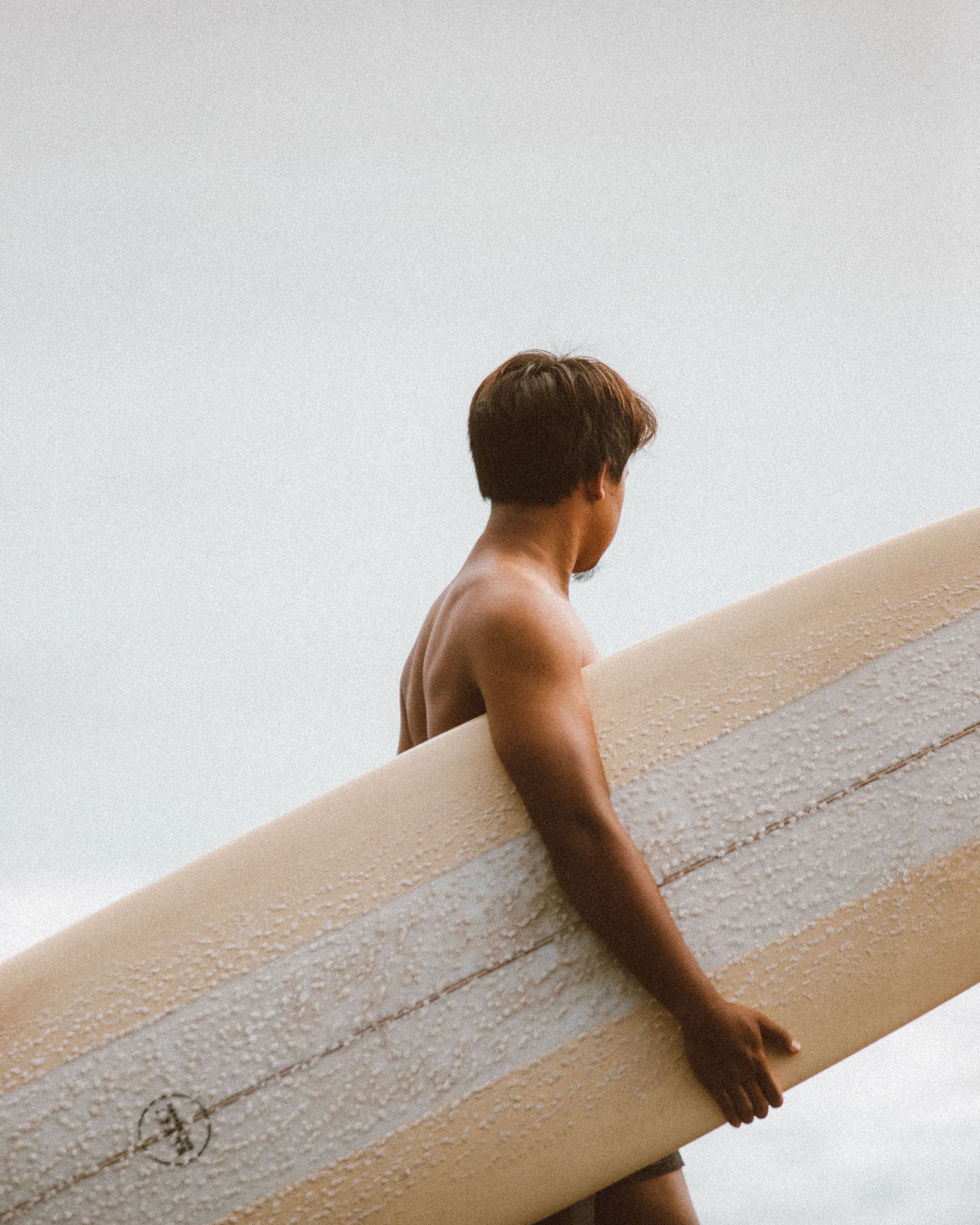 A surfer carrying a longboard along the shore on a bright day, ready to catch the waves.
