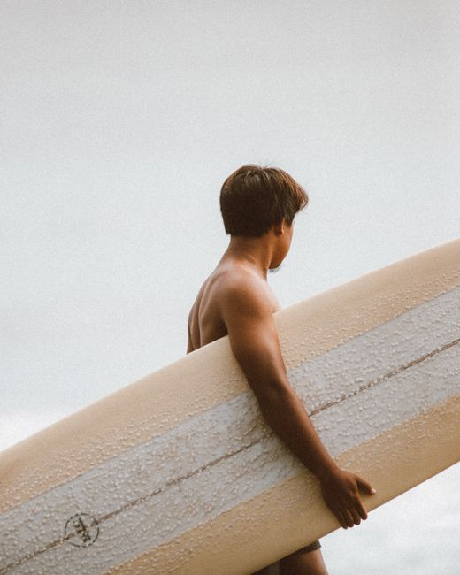 A surfer carrying a longboard along the shore on a bright day, ready to catch the waves.